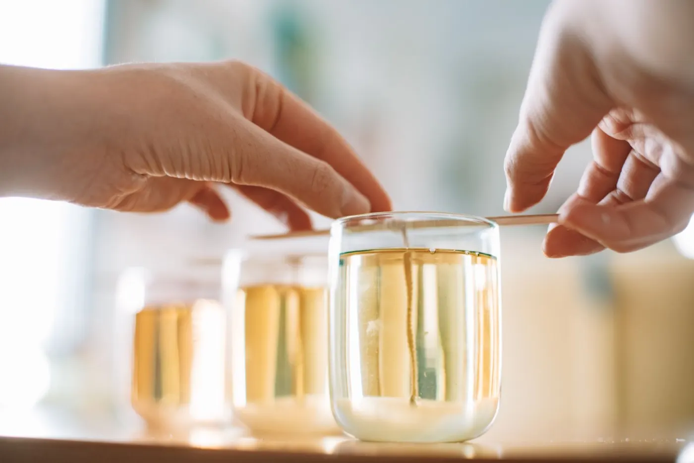 Hands setting a wick in warm wax while glasses of poured candles rest nearby.