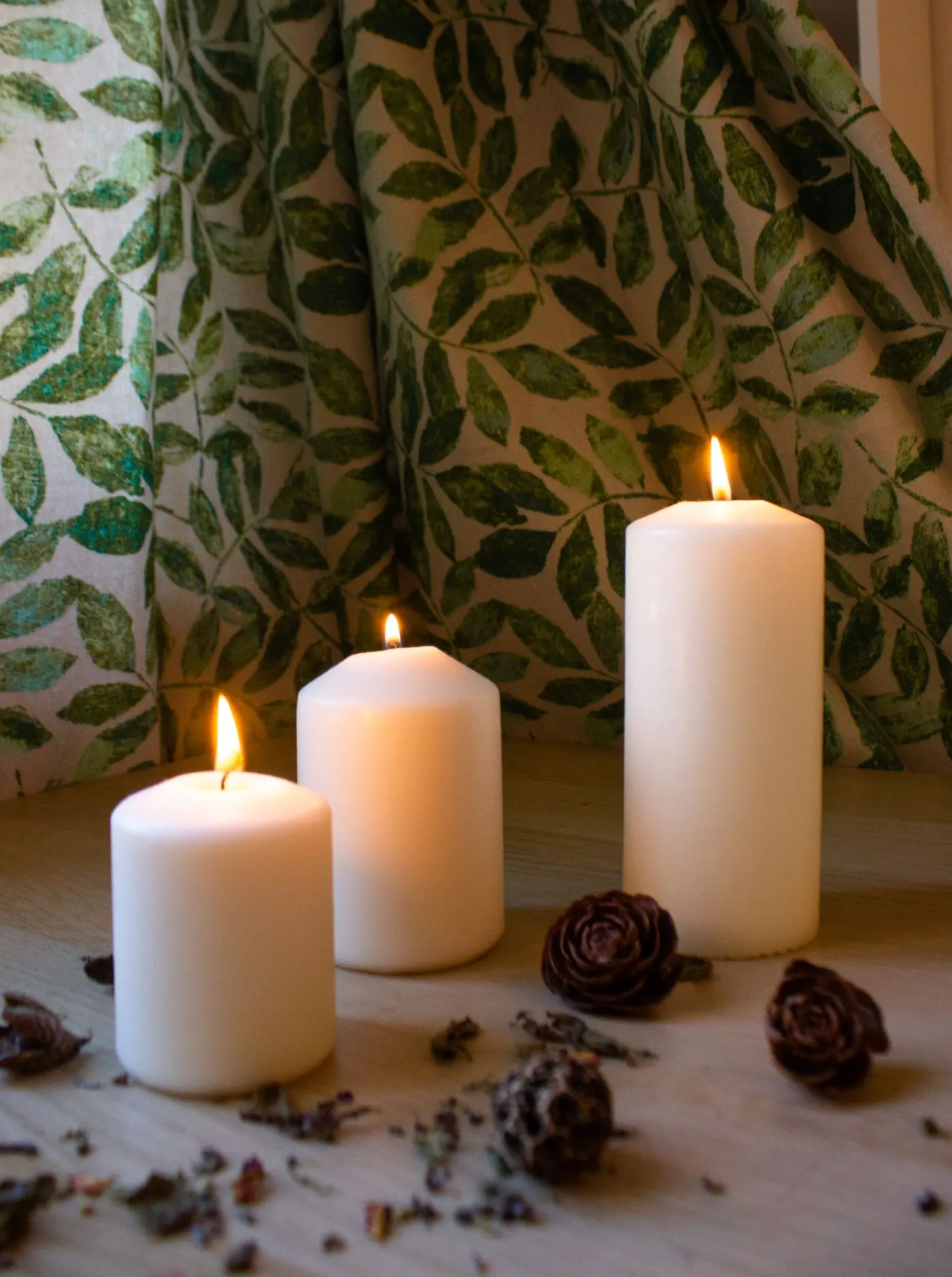 Three lit white pillar candles on a wooden surface with dried botanicals and a leafy curtain.