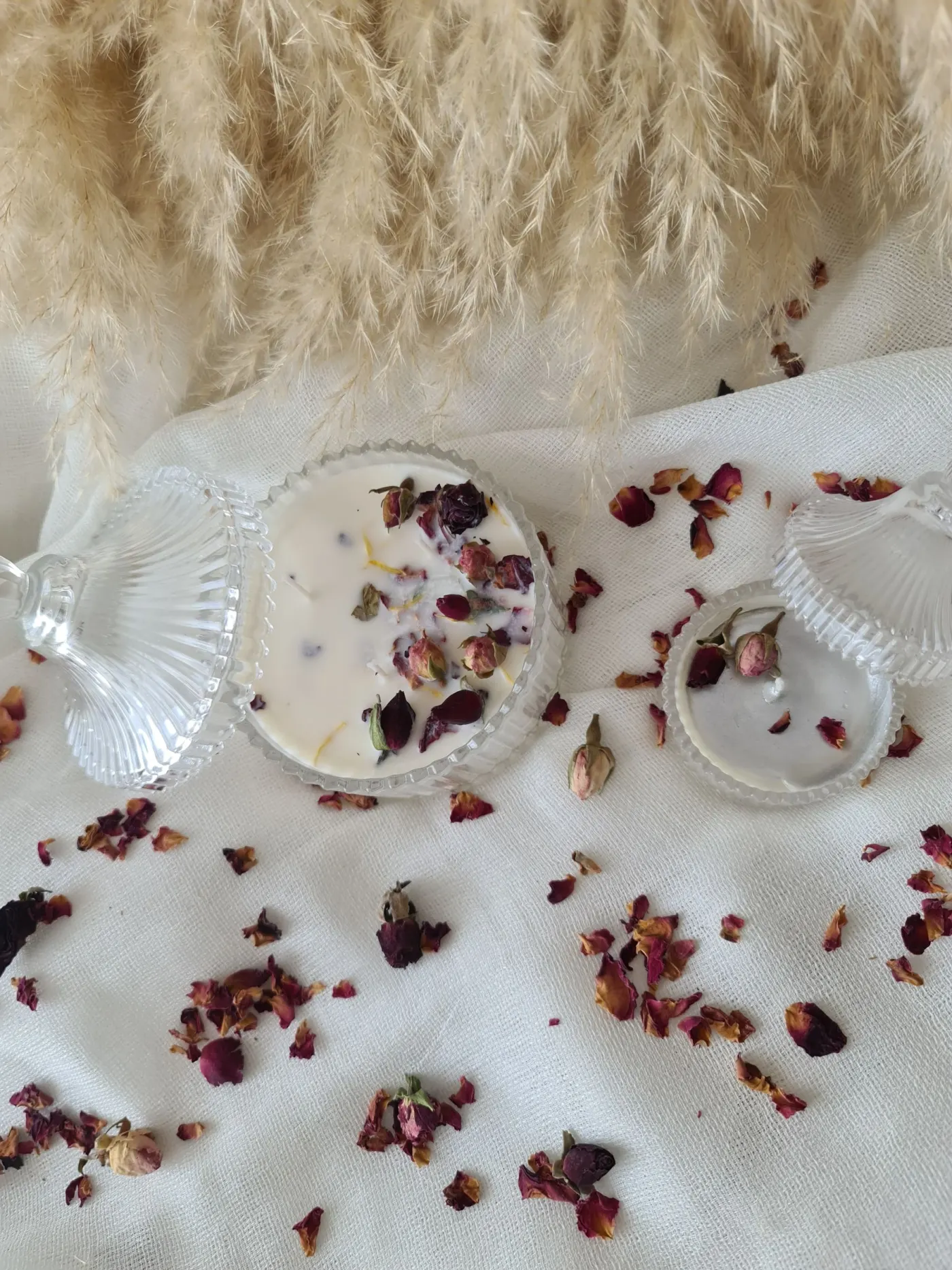 Handmade candles topped with dried rosebuds in glass dishes, arranged on ivory linen with pampas grass.