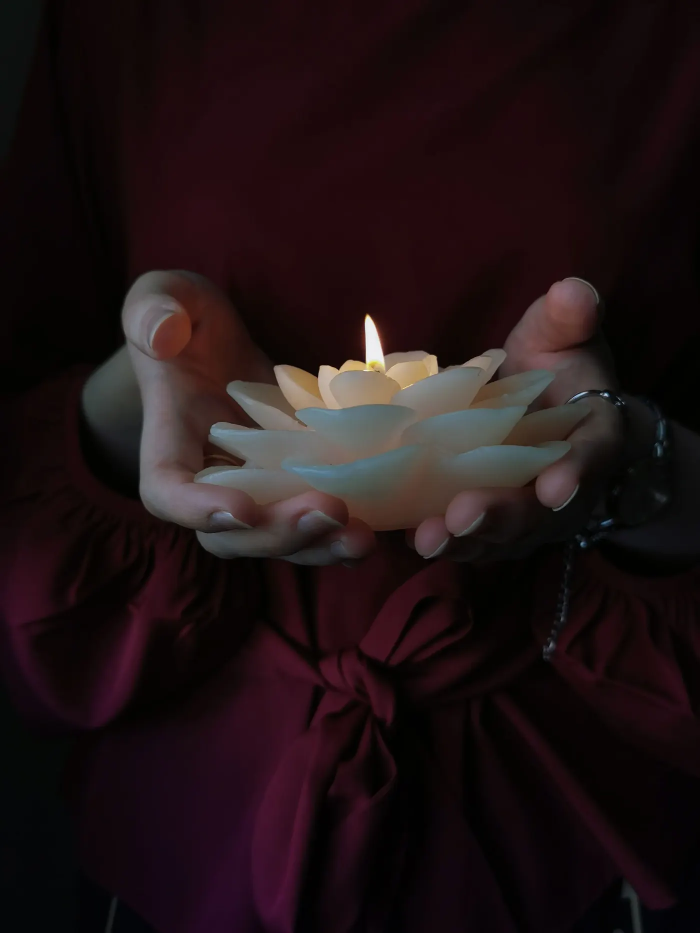A lit lotus-shaped candle held gently in two hands against deep burgundy fabric.