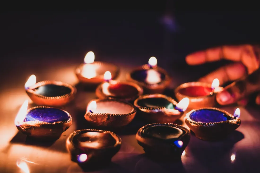 A cluster of colourful clay diyas, several of them lit, on a dark surface.