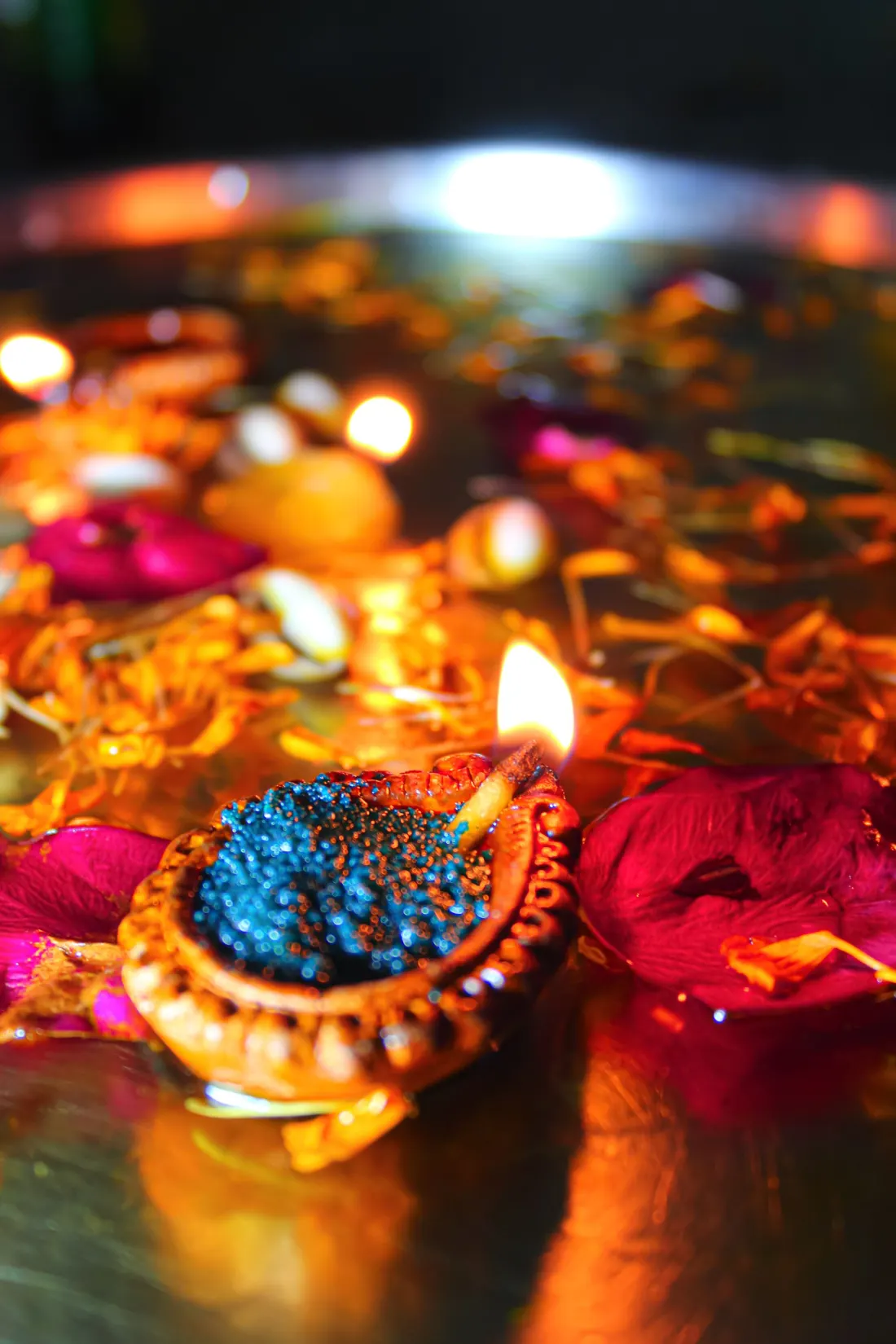 A lit decorative diya with blue detail and rose petals on a brass tray.