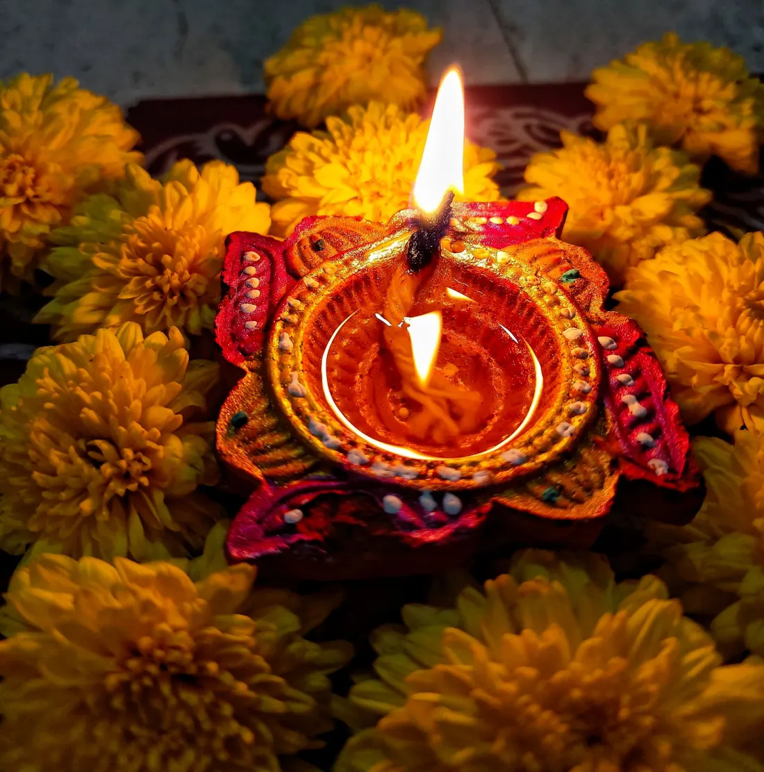 A lit red and gold hand-painted diya surrounded by yellow marigolds.