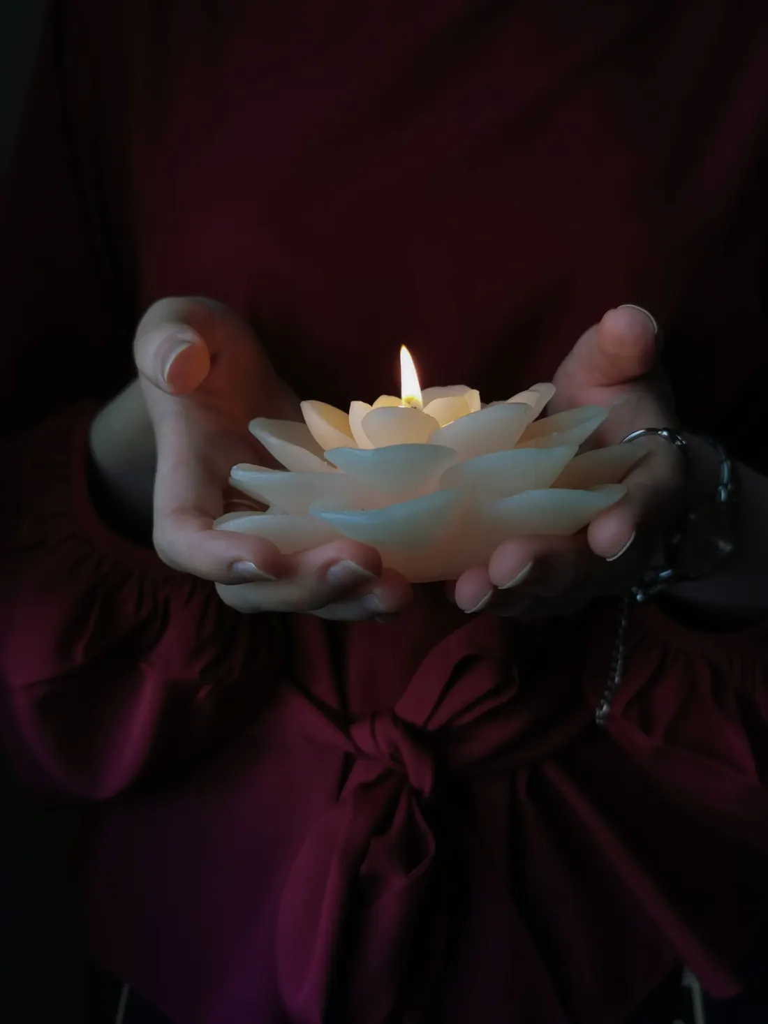 A lit lotus-shaped candle cupped in two hands against burgundy fabric.