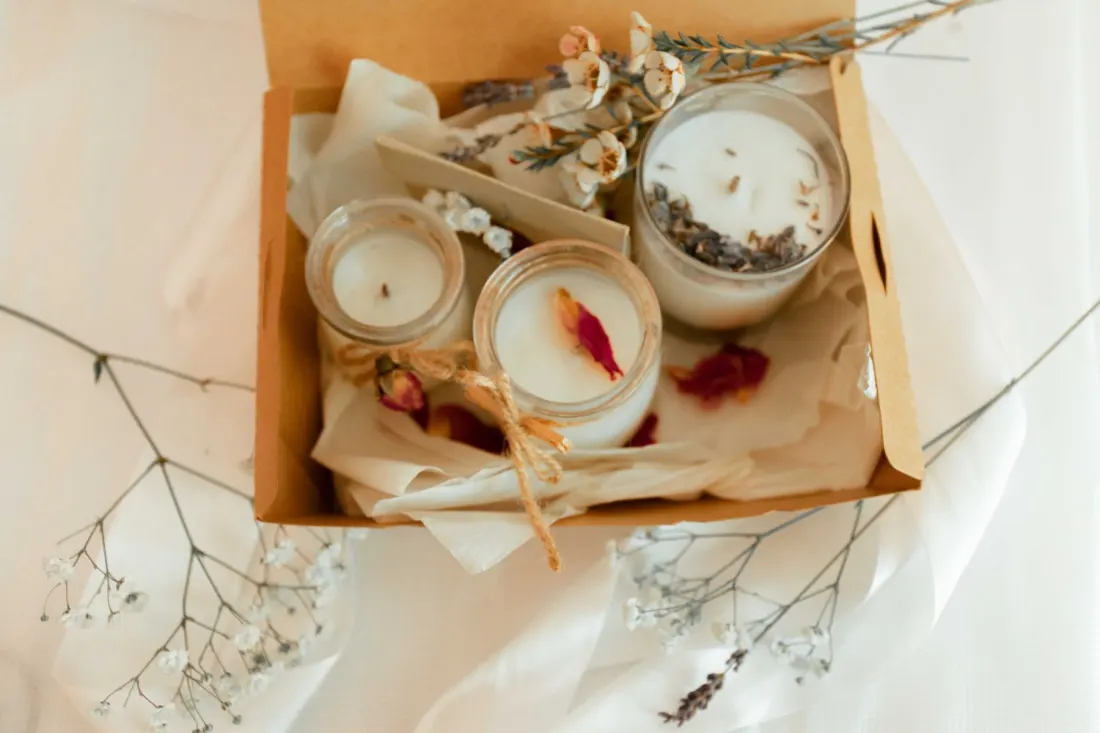 An open kraft gift box holding three candles, tissue, dried flowers, and twine.