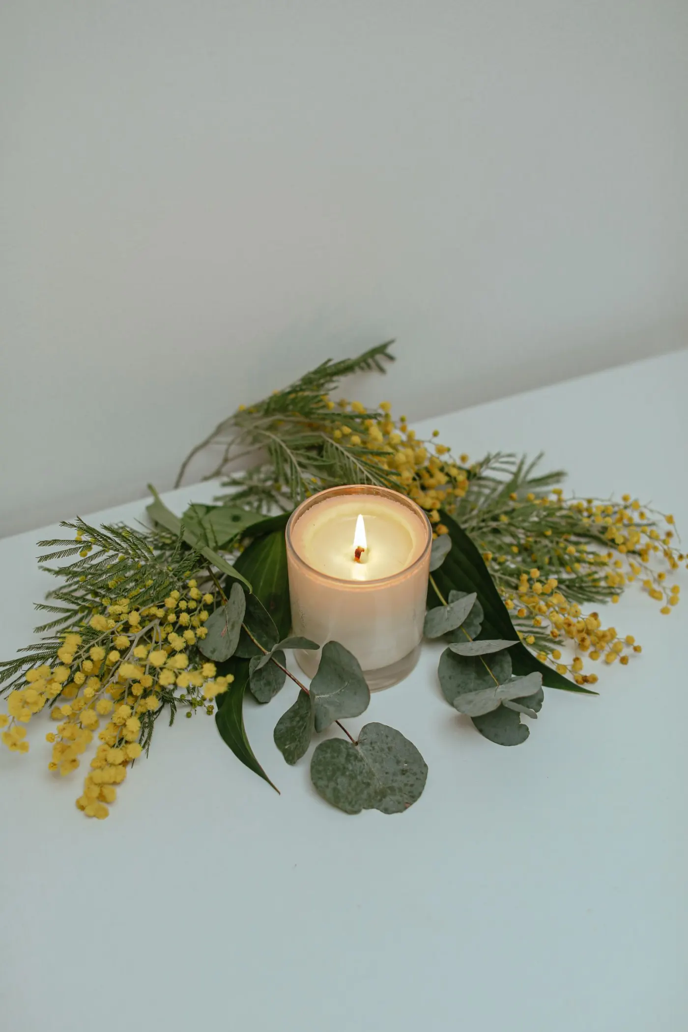 A lit candle in a clear glass, circled by yellow mimosa and eucalyptus on a white table.