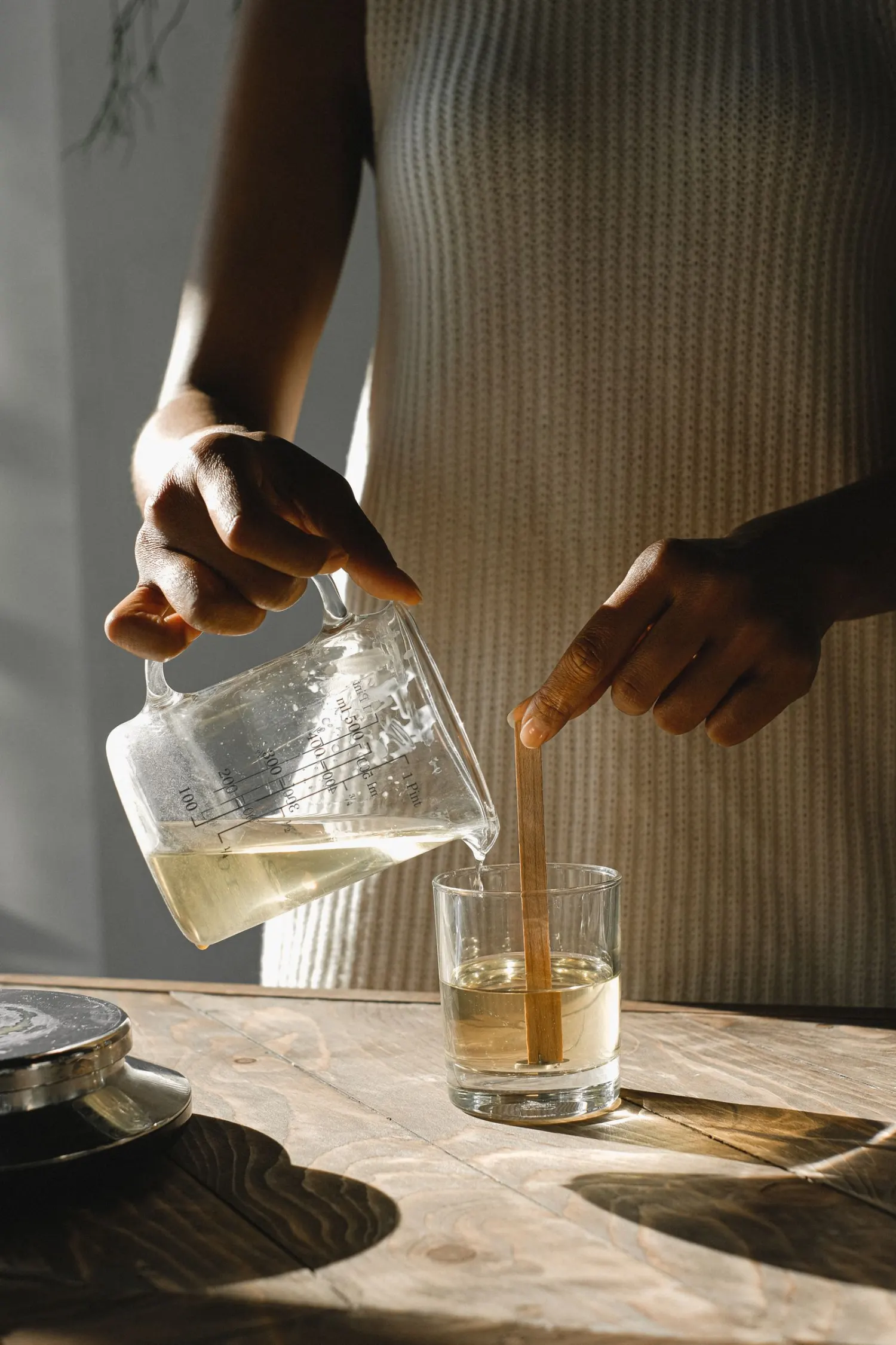A person pouring melted wax into a glass while steadying the wick, in warm window light.
